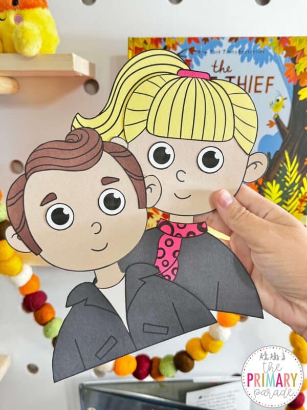 Hand holding retro boy and girl paper crafts with brown and blonde hair, black outfits, and a pink scarf.