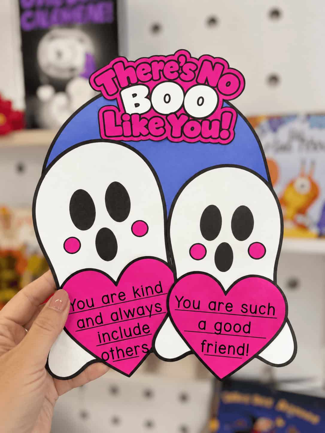 Halloween ghost kindness craft featuring two white ghosts holding pink hearts with friendship compliments, displayed in front of a classroom pegboard.