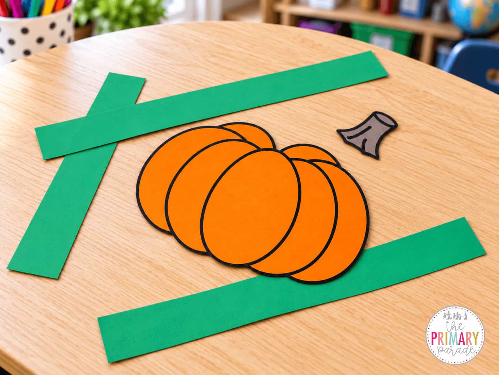 Orange paper pumpkin, separate brown stem, and three green headband strips scattered on a teacher’s desk before assembly.